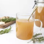Comment faire un Bouillon d'Os : Recettes, Bienfaits et Conseils Essentiels Verre de bouillon d'os avec une cuillère et un bocal en arrière plan. Au ond il y a de l'ail et des herbes sur une plache en bois