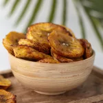 Délicieuse recette de banane plantain au four Banane plantain au four en rondelles dans une coupelle en bois avec une feuille de palmier en arrière plan